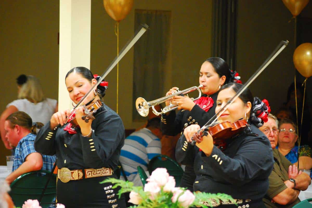 Tri Mariachi devojke sviraju različite instrumente. Jedna devojka svira trubu, dok druge dve sviraju violinu.
